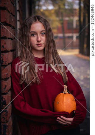 Teen girl in a red sweater holds a pumpkin while leaning against an old brick wall with autumn leaves on the ground in the Timiryazevs Academy, Moscow 132462946