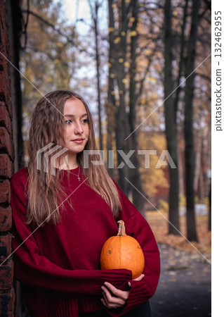 Teen girl in a red sweater holds a pumpkin while leaning against an old brick wall with autumn leaves on the ground in the Timiryazevs Academy, Moscow Teen girl in a red sweater holds a pumpkin while leaning against an old brick wall with autumn leaves on the ground in the Timiryazevs Academy, Moscow 132462955