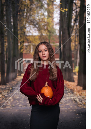 Teen girl in a red sweater holds a pumpkin while standing on an autumn alley surrounded by trees with golden leaves 132462959
