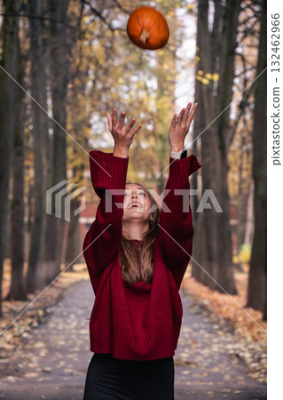 Teen girl in a red sweater throws a pumpkin into the air in an autumn park, surrounded by tall trees and fallen leaves Teen girl in a red sweater throws a pumpkin into the air in an autumn park, surrounded by tall trees and fallen leaves 132462966