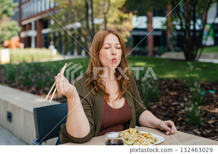 Plus size woman having lunch outdoors, eating noodles. 132463029