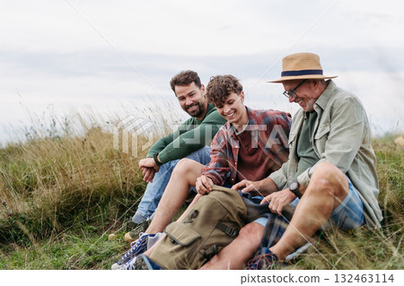 Grandfather, father and teen boy hiking togeter in nature, resting on meadow. 132463114