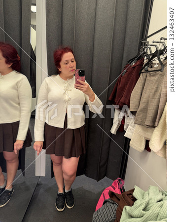 Woman in white cardigan adjusting outfit in fitting room. Style exploration, mirror reflection, and fashion decision moment. 132463407