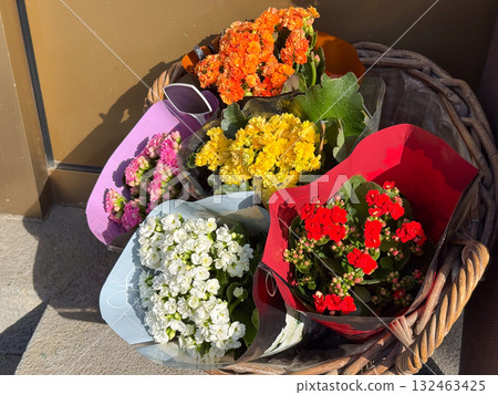 Colorful flower bouquets in paper wraps arranged in a wicker basket outdoors. Bright sunlight highlighting red, yellow, white, orange, and pink blossoms with green leaves. 132463425