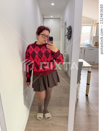 Woman in red argyle sweater and brown skirt taking mirror selfie at home. Cozy outfit, warm light, and casual autumn style. Woman in red argyle sweater and brown skirt taking mirror selfie at home. Cozy outfit, warm light, and casual autumn style. 132463680