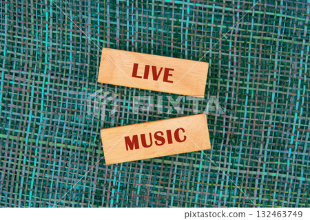 Two wooden blocks spell LIVE MUSIC. The blocks rest on a textured, woven turquoise backdrop. It evokes feelings of fun, excitement, and the joy of experiencing musical performances 132463749