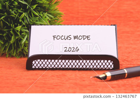 A sleek name card holder displays the words Focus Mode and the year 2026 against an orange backdrop, complemented by a decorative green plant and a classic fountain pen A sleek name card holder displays the words Focus Mode and the year 2026 against an orange backdrop, complemented by a decorative green plant and a classic fountain pen 132463767
