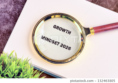 A magnifying glass focuses attention on Growth Mindset 2026 printed on paper. It rests atop a notebook, with a small green plant nearby, signifying forward looking and learning 132463805