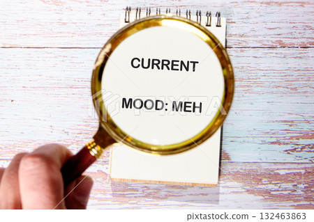 Person holds a magnifying glass over a spiral notepad. The notepad displays the phrase CURRENT MOOD MEH. The notebook sits on a weathered wood surface Person holds a magnifying glass over a spiral notepad. The notepad displays the phrase CURRENT MOOD MEH. The notebook sits on a weathered wood surface 132463863