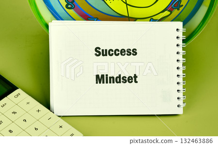 On a vibrant green surface, a notepad displays the words success mindset alongside a calculator and colorful background. This setup invites reflections on goals and achievements 132463886