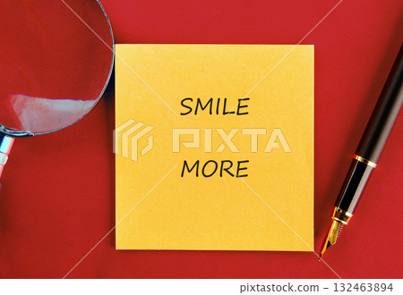 On a red background, a yellow sticky note with SMILE MORE is beside a pen and magnifying glass. A moment of inspiration or reflection on adding positivity to life 132463894
