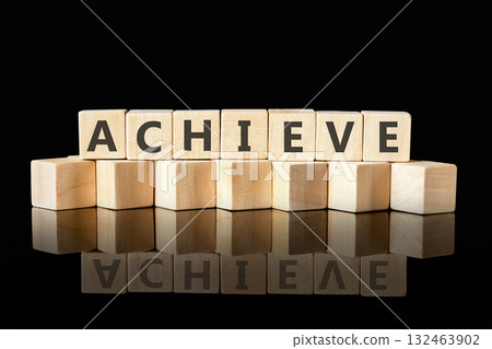 A series of wooden blocks are arranged to spell the word achieve. The blocks reflect light in a dark setting, symbolizing ambition and the pursuit of goals in a simple yet powerful way A series of wooden blocks are arranged to spell the word achieve. The blocks reflect light in a dark setting, symbolizing ambition and the pursuit of goals in a simple yet powerful way 132463902