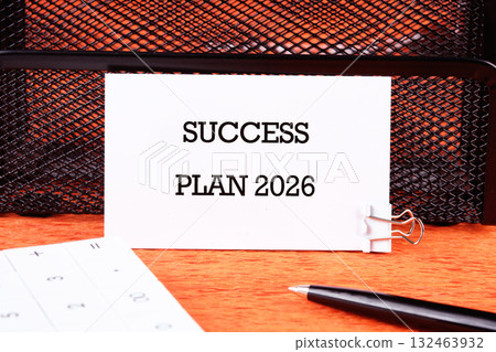 A well-organized workspace features a card reading Success Plan 2026, accompanied by a calculator and pen on an orange surface, highlighting future goals and aspirations A well-organized workspace features a card reading Success Plan 2026, accompanied by a calculator and pen on an orange surface, highlighting future goals and aspirations 132463932