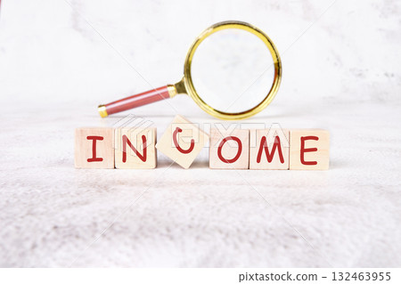 Blocks spelling out income sit on a soft surface, while a magnifying glass emphasizes the word, inviting viewers to explore financial concepts and opportunities in depth 132463955