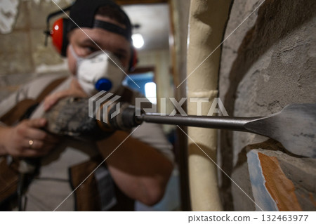 A construction worker using a hammer drill to remove old plaster from an interior wall during a home renovation is performing demolition work while wearing personal protective equipment. A construction worker using a hammer drill to remove old plaster from an interior wall during a home renovation is performing demolition work while wearing personal protective equipment. 132463977