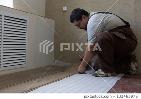 Man kneeling indoors installing laminate planks over white underlayment, carefully aligning floorboards during a residential renovation or diy flooring project in a living space. 132463979
