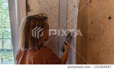 A child uses a putty knife to remove old patterned wallpaper during a home renovation. The child learns practical home improvement skills by helping to update a room in the family home. 132464047
