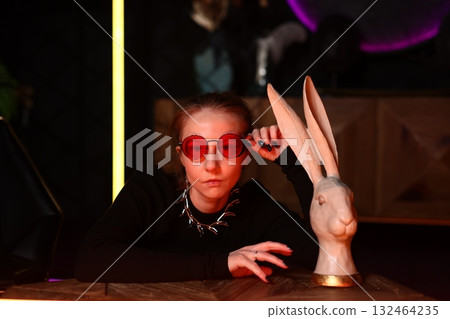 stylish woman wearing red glasses inspects and smiles at rabbit on counter in retro studio setting stylish woman wearing red glasses inspects and smiles at rabbit on counter in retro studio setting 132464235