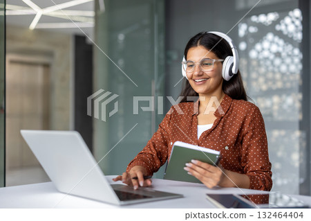 Young woman wearing headphones and glasses, happily engaging in online education or remote work, using her laptop and holding a tablet in a modern office environment Young woman wearing headphones and glasses, happily engaging in online education or remote work, using her laptop and holding a tablet in a modern office environment 132464404