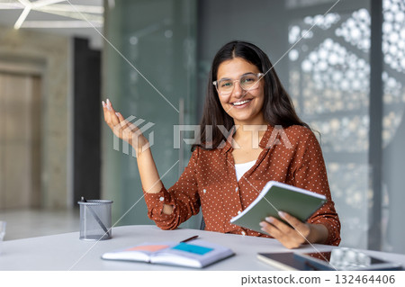 Indian businesswoman smiling at her office desk, holding a notebook and gesturing confidently while leading a meeting or presentation, mentoring colleagues and discussing strategy 132464406