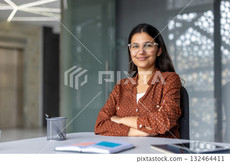 Young indian businesswoman sitting at an office desk, smiling confidently while wearing glasses and brown polka dot shirt, conveying professionalism and positive attitude Young indian businesswoman sitting at an office desk, smiling confidently while wearing glasses and brown polka dot shirt, conveying professionalism and positive attitude 132464411