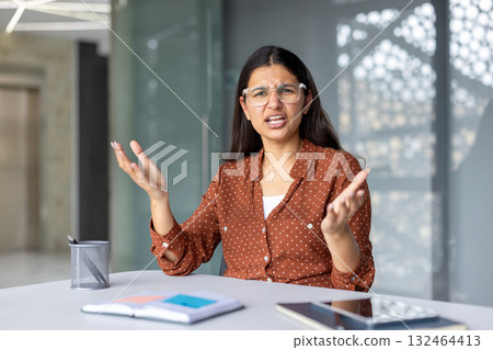Young businesswoman wearing glasses sits at her desk, engaging in a video call. Raising her hands, she passionately argues while navigating a digital discussion on her laptop 132464413