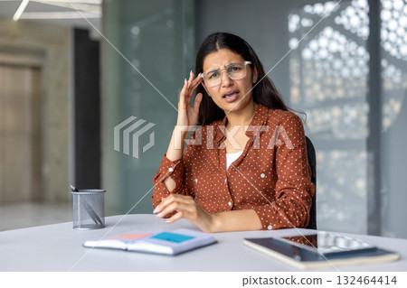 Indian businesswoman in eyeglasses feeling severe headache or migraine, suffering from pain and pressure, needing medical attention while sitting at her office desk 132464414