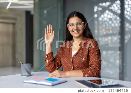 Young indian business woman sitting at a desk and raising her hand, smiling while participating in a video call or online meeting in a modern office environment 132464416