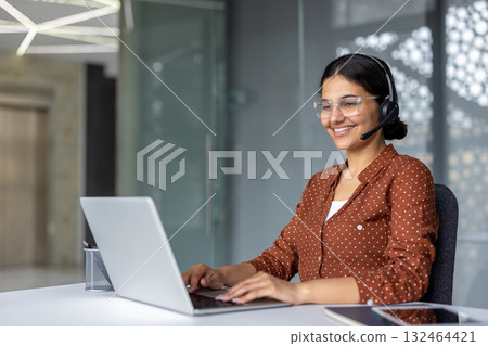 Young woman wearing a headset, typing on a laptop keyboard and smiling while working remotely in a modern office space, exuding confidence and professionalism in her career Young woman wearing a headset, typing on a laptop keyboard and smiling while working remotely in a modern office space, exuding confidence and professionalism in her career 132464421