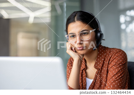 Bored female call center operator with headset is resting her chin on her hand while looking at laptop screen, having a difficult day at work Bored female call center operator with headset is resting her chin on her hand while looking at laptop screen, having a difficult day at work 132464434