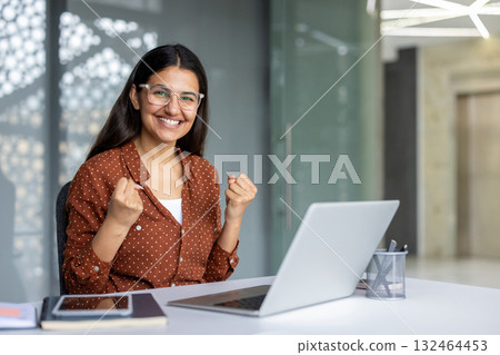 Businesswoman with glasses is sitting at her desk, raising her fists in a gesture of triumph, expressing her excitement and achievement after successfully completing a task on her laptop Businesswoman with glasses is sitting at her desk, raising her fists in a gesture of triumph, expressing her excitement and achievement after successfully completing a task on her laptop 132464453