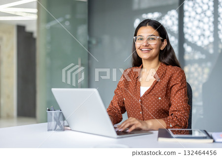 Indian businesswoman smiling confidently. Wearing glasses. And typing on a laptop while sitting at a clean desk in a modern corporate office environment. Representing professionalism and productivity 132464456