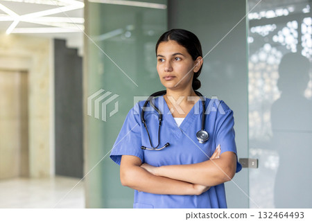 Healthcare professional woman wearing blue scrubs and a stethoscope, standing with arms crossed and looking away, representing dedication and critical thinking in her medical role Healthcare professional woman wearing blue scrubs and a stethoscope, standing with arms crossed and looking away, representing dedication and critical thinking in her medical role 132464493