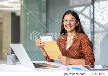 Young indian woman smiling with excitement as she opens a letter at her desk in a modern office, celebrating a job offer or important career news while working on a laptop Young indian woman smiling with excitement as she opens a letter at her desk in a modern office, celebrating a job offer or important career news while working on a laptop 132464552
