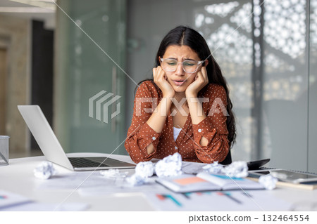 Young woman feeling overwhelmed and frustrated by her work, sitting at a desk with a laptop and crumpled papers, showing signs of stress and a tight deadline Young woman feeling overwhelmed and frustrated by her work, sitting at a desk with a laptop and crumpled papers, showing signs of stress and a tight deadline 132464554