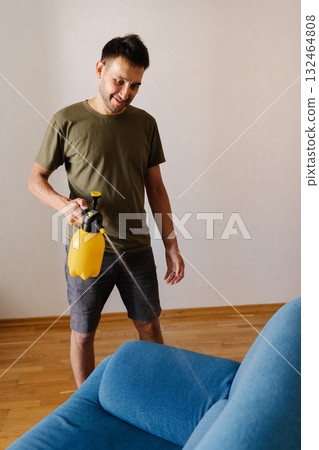 Happy smiling young man spraying water using yellow plastic sprayer on blue sofa in stylish living room, engaging in thorough home cleaning and maintenance. Concept of household cleanliness. Happy smiling young man spraying water using yellow plastic sprayer on blue sofa in stylish living room, engaging in thorough home cleaning and maintenance. Concept of household cleanliness. 132464808