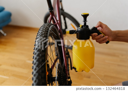 Cyclist cleaning mountain bike wheel with yellow spray bottle, removing dust and dirt from tire, performing detailed maintenance and care inside workshop or home garage, close-up. Cyclist cleaning mountain bike wheel with yellow spray bottle, removing dust and dirt from tire, performing detailed maintenance and care inside workshop or home garage, close-up. 132464818