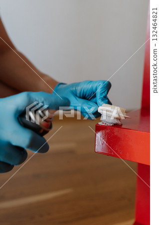 Vertical cropped shot of man wearing blue protective gloves removing adhesive stains, scrubbing red wooden shelf with small brush during detailed cleaning process at home, close-up. 132464821