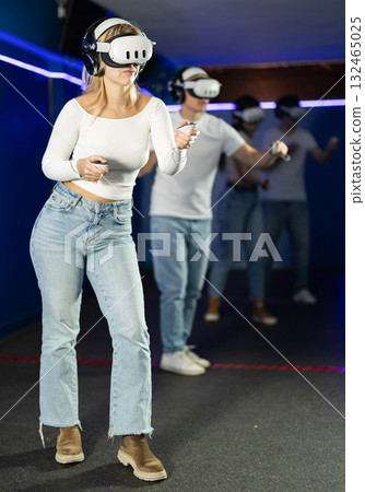 In darkened hall of gaming club, girl in VR helmet takes part in group game 132465025