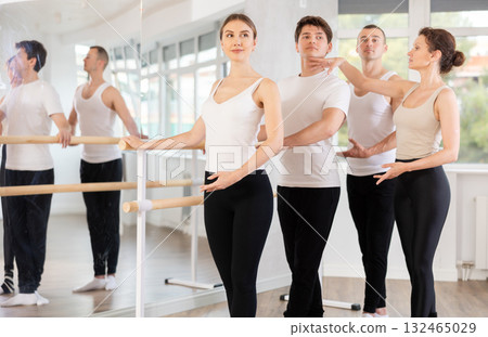 Professional female ballet trainer teaching group of dancers, helps to coordinate movements correctly. Young girl and guys near ballet barre in dancing hall Professional female ballet trainer teaching group of dancers, helps to coordinate movements correctly. Young girl and guys near ballet barre in dancing hall 132465029