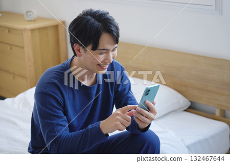 A man in his twenties sitting on a bed and using a smartphone A man in his twenties sitting on a bed and using a smartphone 132467644