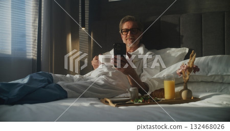 Mature Executive Sits on Hotel Bed, Dressed in Bathrobe, Checking Smartphone While Holding Coffee 132468026