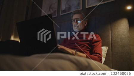 Focused Man Works on Laptop in Dark Hotel Room 132468056