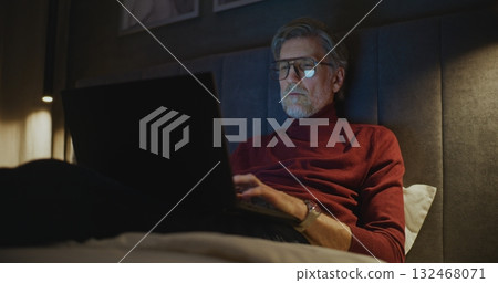 Senior Man With Personal Computer Sits Comfortably on Bed in Softly Lit Modern Hotel Room Senior Man With Personal Computer Sits Comfortably on Bed in Softly Lit Modern Hotel Room 132468071