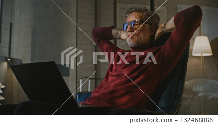 Gray Haired Man Sits by Bed, Working on Laptop in Evening Light of Hotel Room Gray Haired Man Sits by Bed, Working on Laptop in Evening Light of Hotel Room 132468086