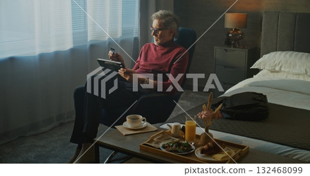 Mature Man in Stylish Hotel Room Enjoys Breakfast in Morning While Browsing Online Store on Tablet 132468099