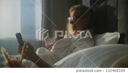 Mature Executive Sits on Hotel Bed, Dressed in Bathrobe, Checking Smartphone While Holding Coffee Mature Executive Sits on Hotel Bed, Dressed in Bathrobe, Checking Smartphone While Holding Coffee 132468109