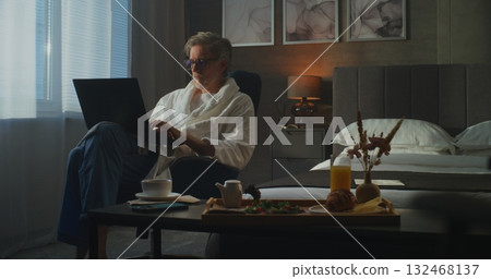 Mature Man in White Robe Works on Laptop in Cozy Hotel Room, With Breakfast and Coffee on Table Mature Man in White Robe Works on Laptop in Cozy Hotel Room, With Breakfast and Coffee on Table 132468137