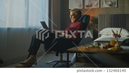 Focused Senior Businessman Works on Laptop in Soft Morning Light of Hotel Room 132468140