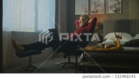 Gray Haired Man Sits Relaxed in Hotel Chair, Typing on Laptop in Morning Light 132468145